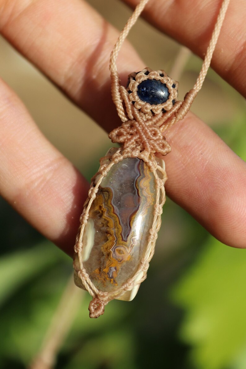 K&ouml;nnte beinhalten: Handgefertigte Makramee-Halskette mit einem polierten, oval geformten Stein mit braunen, gelben und wei&szlig;en Mustern. Der Stein ist in einem beigefarbenen Makramee-Gewebe gefasst, mit einem kleinen blauen Stein-Akzent oben.