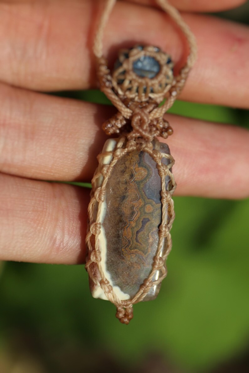 K&ouml;nnte beinhalten: Eine handgefertigte Anh&auml;nger Halskette mit einem braunen und cremefarbenen Stein mit komplizierten Mustern. Der Stein ist in einer Kordel im Makramee-Stil gefasst, mit einem kleinen blauen Steinakzent oben. Die Halskette ist an einer beigefarbenen Kordel aufgeh&auml;ngt.