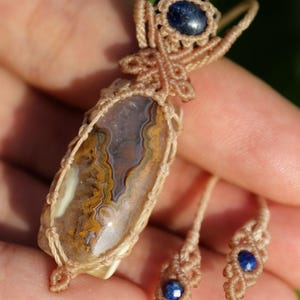 May include: A handcrafted macrame pendant necklace and matching earrings. The pendant features a polished, oval-shaped stone with brown, yellow, and white patterns, encased in beige macrame. The earrings each have a small blue stone.