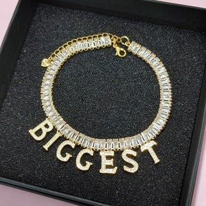May include: Gold-toned bracelet with baguette-cut clear crystals and the word "BIGGEST" spelled out in individual letter charms. The bracelet is displayed in a black box on a pink surface.
