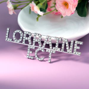 May include: A silver-toned brooch spelling "LORRAINE ECT" in capital letters, encrusted with clear rhinestones. The brooch is set against a soft purple background, with pink flowers and a white dish in the background.