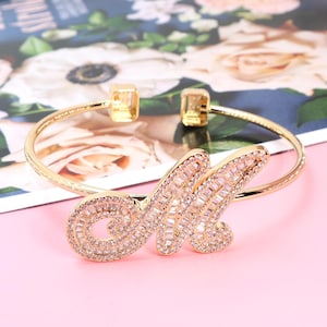Puede incluir: Brazalete dorado con la letra "M" incrustada con cristales transparentes. El brazalete tiene un diseño sencillo y abierto con extremos cuadrados. El fondo es un estampado floral sobre una superficie rosa.