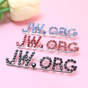 May include: Three brooches spelling "JW.ORG" in blue, red, and black rhinestones. The brooches are on a pink background, with a white flower in the upper left corner.