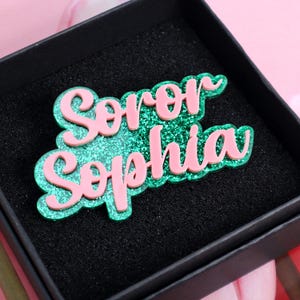 Puede incluir: Un broche rosa y verde brillante con las palabras "Soror Sophia" en fuente cursiva. El broche está en una caja negra, con un fondo rosa. El broche es un accesorio decorativo.