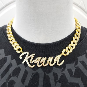 Könnte beinhalten: Eine goldene Kettenkette mit einem großen, kursiv geschriebenen Namenspannen, auf dem "Kiara" in weißen Strasssteinen steht.