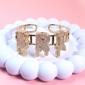 May include: A gold-toned cuff bracelet with the word "BRI" spelled out in sparkling, gem-studded letters. The letters have a dripping effect. The bracelet is displayed on a white, circular, beaded platform against a pink background.