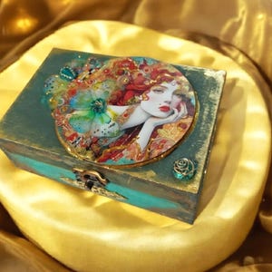 May include: A teal wooden box with a decorative lid featuring a woman's portrait with red hair and a butterfly. The box has gold accents, a metal clasp, and a small teal rose embellishment. The box is sitting on a gold satin surface.