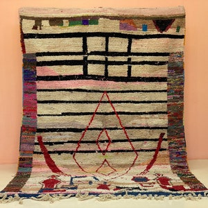 Puede incluir: Una alfombra marroquí colorida con un diseño geométrico que presenta rayas negras, una forma de diamante rosa y blanco y acentos rojos y azules. La alfombra tiene un borde de flecos.