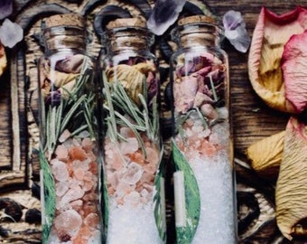 May include: Three glass bottles with cork tops filled with pink Himalayan salt, white sea salt, and black lava salt. The bottles are decorated with dried flowers, herbs, and leaves.