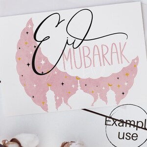 May include: A pink and white card with the text "Eid Mubarak" in black cursive script. The background features a pink crescent moon with gold stars and white silhouettes of mosques.