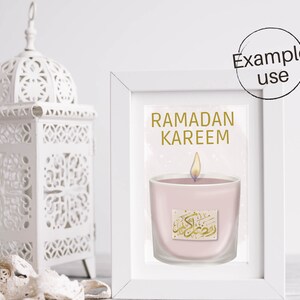 May include: A white frame with a gold inscription that reads "Ramadan Kareem". Inside the frame is a pink candle with a gold inscription in Arabic. The candle is lit and has a white flame.