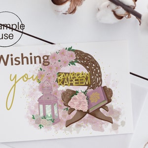 May include: A greeting card with a watercolor background featuring a wreath of pink flowers, a lantern, a book, and a sign that reads "Ramadan Kareem". The text "Wishing you" is written in gold script.