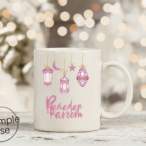 Op de afbeelding: Witte keramische mok met roze aquarel lantaarns, een maan en een ster, opgehangen aan gouden touwtjes. De woorden "Ramadan Kareem" staan in roze letters op de mok.