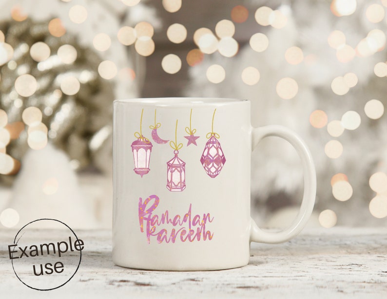 Op de afbeelding: Witte keramische mok met roze aquarel lantaarns, een maan en een ster, opgehangen aan gouden touwtjes. De woorden "Ramadan Kareem" staan in roze letters op de mok.