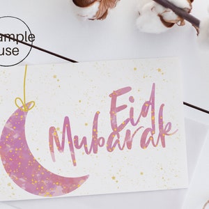 Op de afbeelding: Een wenskaart met de tekst "Eid Mubarak" in roze script, vergezeld van een roze halve maan met gouden sterren. De kaart heeft een witte achtergrond met gouden spikkels en ligt op een witte ondergrond.