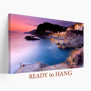 Puede incluir: Una colorida puesta de sol sobre una ciudad costera con una costa rocosa y un pequeño muelle. El agua es de un azul profundo y el cielo es una mezcla de rosa, morado y naranja.