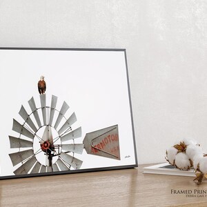 May include: A framed wall art print featuring a photograph of a bald eagle perched atop a metal windmill. The windmill blades are silver and circular, with a tail vane displaying the red text AERMOTOR USA. The frame is thin and black.
