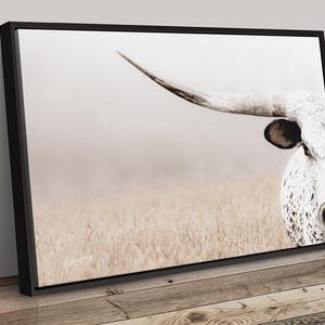 May include: A rectangular wall art canvas featuring a close-up of a white longhorn cow with dark spots, a prominent curved horn, and a dark ear. The cow is positioned on the right side against a soft, light-coloured, hazy field background.
