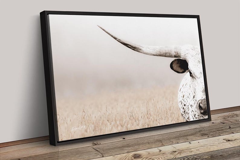 May include: A rectangular wall art canvas featuring a close-up of a white longhorn cow with dark spots, a prominent curved horn, and a dark ear. The cow is positioned on the right side against a soft, light-coloured, hazy field background.