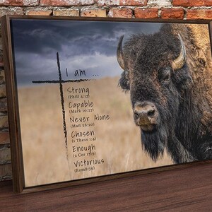 May include: A rectangular framed wall art print featuring a large brown bison on the right side and a black cross on the left. The text reads: I am... Strong (Phil 4:13), Capable (Mark 10:27), Never Alone (Matt 28:20), Chosen (1 Thess 1:4), Enough (2 Cor 12:9), Victorious (Rom 8:39).