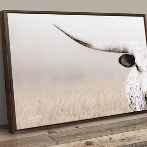 May include: A rectangular wall art print featuring a close-up of a white longhorn cow with dark spots, a prominent curved horn, and a black ear. The animal is positioned on the right side against a soft, light-coloured, hazy field background.