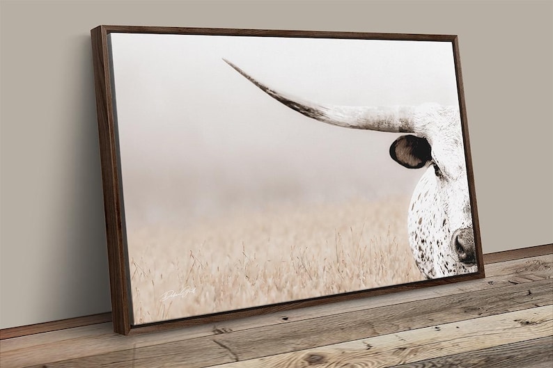 May include: A rectangular wall art print featuring a close-up of a white longhorn cow with dark spots, a prominent curved horn, and a black ear. The animal is positioned on the right side against a soft, light-coloured, hazy field background.