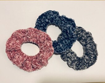 May include: Three scrunchies in different colors. One is pink, one is gray, and one is a darker shade of gray. They are all made of a soft, fuzzy material.