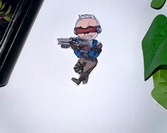Può includere: Un adesivo con un'illustrazione di un personaggio dei cartoni animati in uniforme blu e marrone, che tiene in mano una pistola. Il personaggio ha i capelli bianchi e occhiali rossi. L'adesivo è su una superficie bianca.