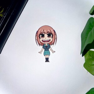 May include: A sticker featuring a chibi-style anime character with short, light brown hair and large brown eyes. The character wears a teal top and skirt with black shoes, smiling with an open mouth. The sticker is on a white background.