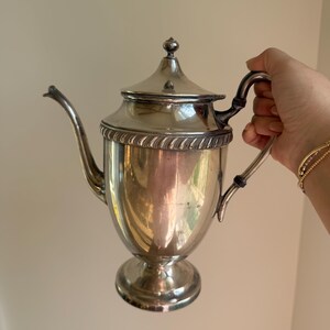 May include: A silver-colored teapot with a curved spout, a lid, and a handle. The teapot has a decorative rope-like design around the top and sits on a pedestal base. The teapot is held by a person.