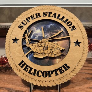 May include: A wooden plaque with the words "SUPER STALLION HELICOPTER" and a helicopter illustration. The plaque has a circular design with a rope-like border and star accents. The helicopter is set against a background of a stormy sky.