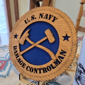 May include: Wooden plaque with a blue center featuring crossed axe and hammer symbols, and the text "U.S. NAVY DAMAGE CONTROLMAN" in a circular design. The plaque has a rope-like border and star cutouts.