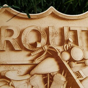 May include: A wooden sign with the word "ROUTE" carved into it. The sign is shaped like a shield and features a motorcycle with a headlight and handlebars.