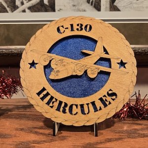 May include: Wooden C-130 Hercules aircraft plaque. The round plaque features a detailed outline of the aircraft, the text "C-130" and "HERCULES", and two stars. The background is blue, and the outer ring is a light brown color.