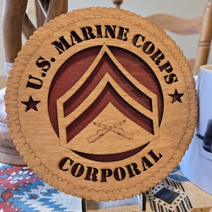 May include: A wooden plaque with the text "U.S. MARINE CORPS CORPORAL" in a circular design. The center features a Corporal rank insignia and crossed rifles. The plaque has a rope-like border and is made of light brown wood with a red center.