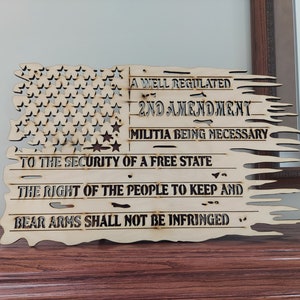 May include: A wooden American flag with a distressed look, featuring the text "A well regulated Militia being necessary to the security of a free state, the right of the people to keep and bear arms shall not be infringed."