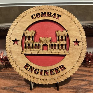 May include: Wooden Combat Engineer plaque with a red center and a decorative border. The design features a castle-like structure and the words "COMBAT ENGINEER" cut out. Two red stars are on either side of the castle.