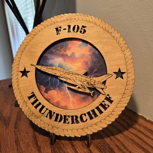 May include: A wooden F-105 Thunderchief plaque. The circular plaque features a detailed laser-cut image of the aircraft, set against a colorful sky background. The text "F-105" and "Thunderchief" are prominently displayed.