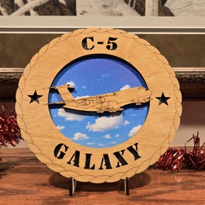 May include: Wooden C-5 Galaxy aircraft plaque. The round plaque features a laser-cut design of the aircraft against a blue sky with clouds. The text "C-5" and "GALAXY" are also laser-cut into the wood. Two black stars are cut into the wooden frame.