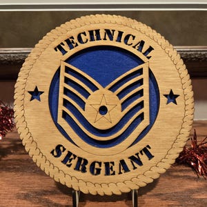 May include: A wooden plaque with the words "TECHNICAL SERGEANT" cut out. The center features a blue background with a military insignia. Two stars flank the insignia. The plaque is round with a rope-like border.