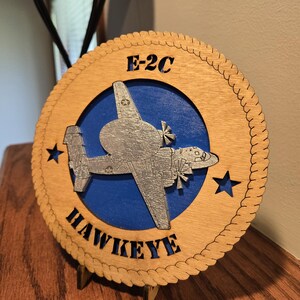 May include: Wooden plaque featuring an E-2C Hawkeye aircraft design. The round plaque has a blue center with the aircraft, surrounded by a wooden border with the text "E-2C HAWKEYE". Two stars are on the sides. The plaque is on a wooden stand.