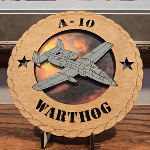 May include: A wooden plaque featuring a gray A-10 Warthog aircraft design. The text "A-10 WARTHOG" is displayed in black lettering. The plaque has a decorative edge and includes two black star accents. The background shows a sunset.
