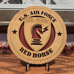 May include: A wooden coaster with the text "U.S. AIR FORCE RED HORSE" in a circular design. The coaster features a red horse emblem and two stars. The coaster is displayed on a small stand.