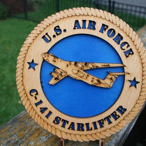 May include: A wooden round plaque with a blue background and a white silhouette of a C-141 Starlifter aircraft. The plaque has a rope-like border and the text "U.S. AIR FORCE C-141 STARLIFTER" around the edge.