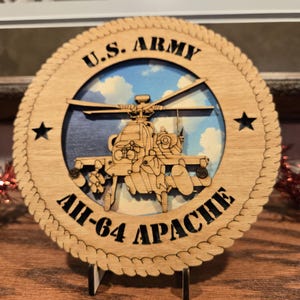 May include: A wooden round plaque featuring a laser-cut design of an AH-64 Apache helicopter. The plaque has the text "U.S. ARMY" at the top and "AH-64 APACHE" at the bottom. The background shows a blue sky with clouds. Two black stars are on the sides.