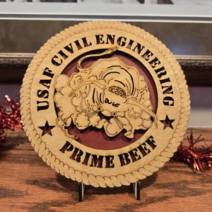 May include: A round wooden plaque with the text "USAF CIVIL ENGINEERING PRIME BEEF" in a circular design. The center features a detailed illustration of a bull. The plaque is supported by a small stand.