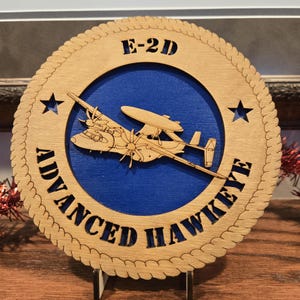 May include: A wooden plaque featuring an E-2D Advanced Hawkeye aircraft design. The design is laser-cut with a light brown finish, set against a blue background. The text "E-2D" and "ADVANCED HAWKEYE" are also cut into the wood, with star accents.