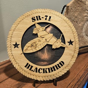 May include: Wooden SR-71 Blackbird plaque. The round plaque features a laser-cut design of the aircraft, the text "SR-71" and "BLACKBIRD", and two stars. The aircraft is set against a dark background with a lightning bolt.