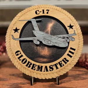 May include: Wooden plaque featuring a gray C-17 Globemaster III aircraft design. The plaque has a circular shape with a rope-like border and the text "C-17 GLOBEMASTER III". Two black stars are on the plaque. The background shows a lightning storm.