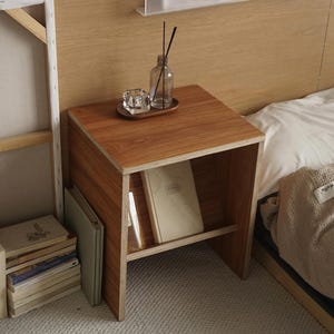 Könnte beinhalten: Ein kleiner Nachttisch aus Holz mit Holzmaserung. Er hat ein Regal und beherbergt Bücher. Eine Glasdiffusorflasche mit Schilf, ein kleiner Glaskerzenhalter und ein Holztablett stehen obenauf. Der Tisch steht neben einem Bett.